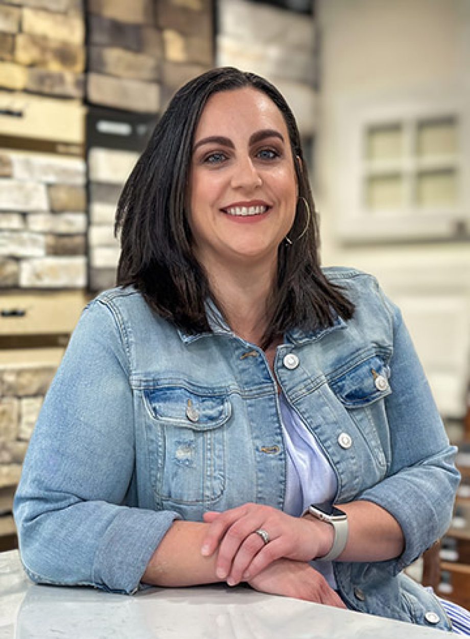 Professional headshot of an S&A Homes team member wearing a light denim jacket, smiling in a design center with cabinetry and finish samples in the background.