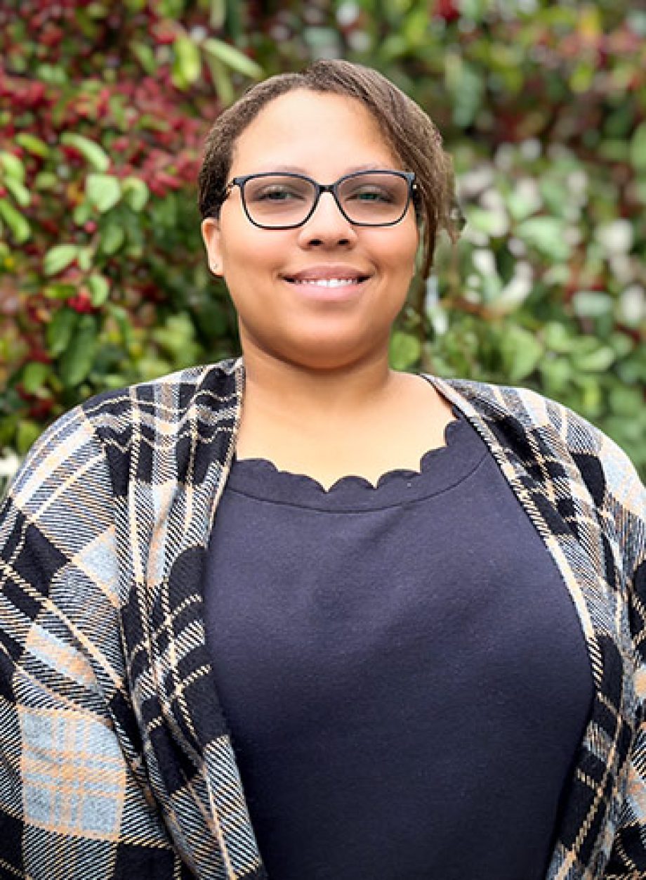 Professional headshot of an S&A Homes team member wearing glasses and a plaid cardigan, smiling outdoors with green foliage in the background.