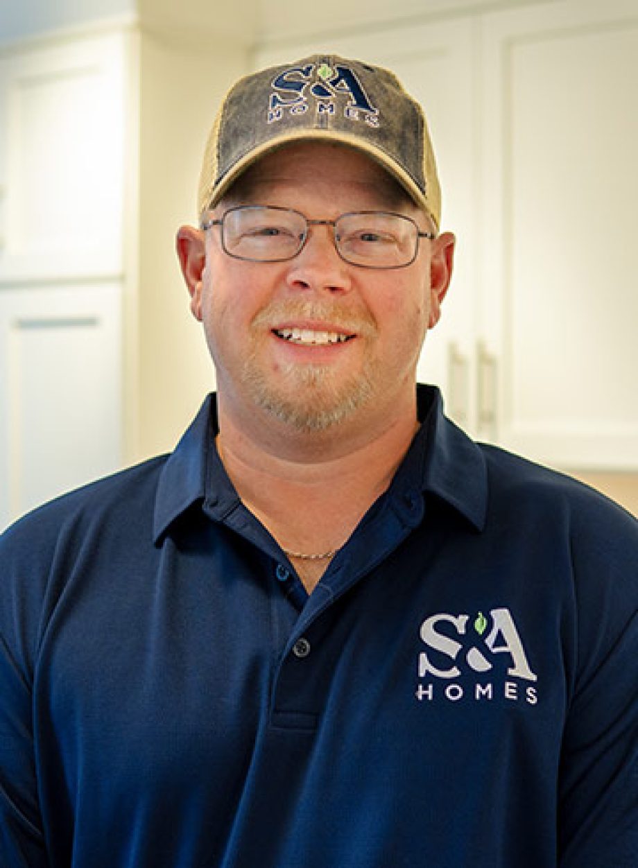 Professional headshot of an S&A Homes team member wearing glasses, an S&A Homes polo shirt, and a baseball cap, smiling in a model home kitchen.