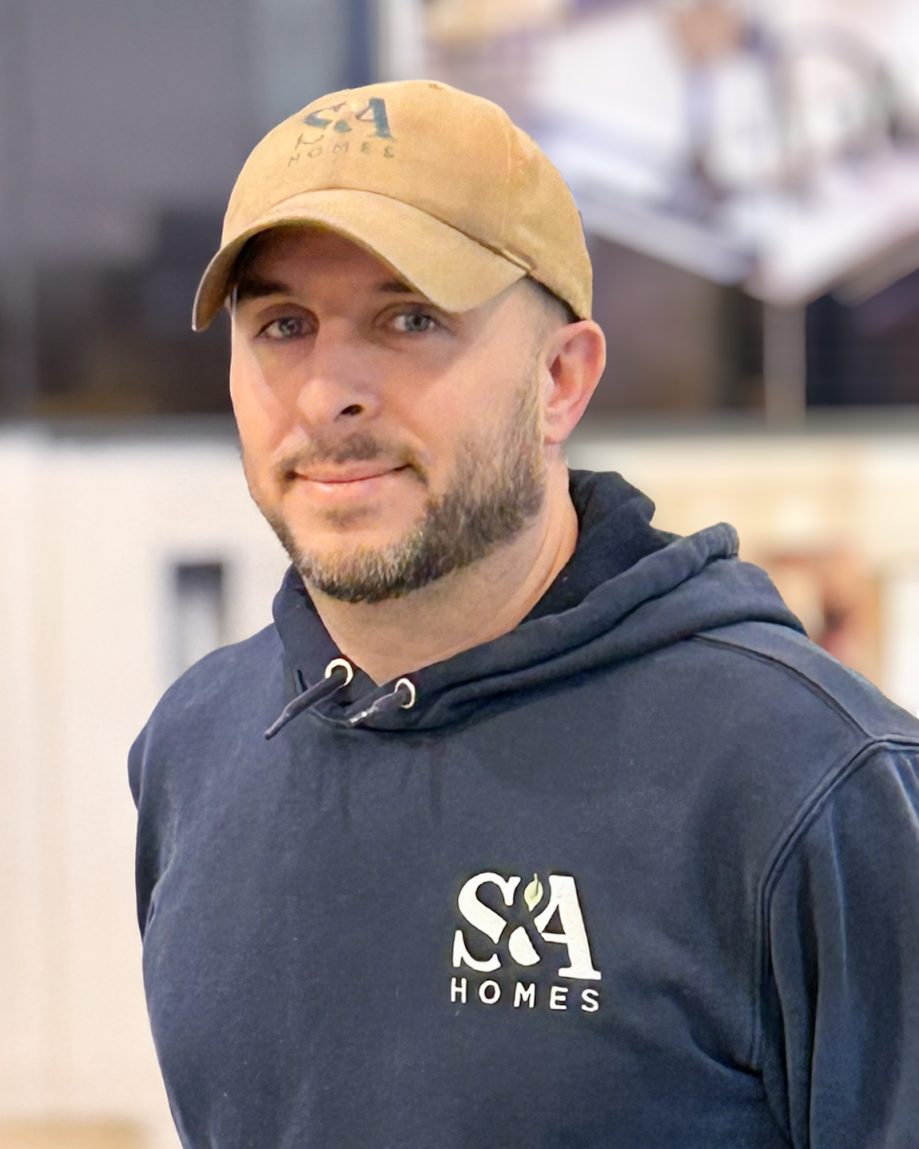 Professional headshot of an S&A Homes team member wearing an S&A Homes hoodie and baseball cap, smiling indoors.