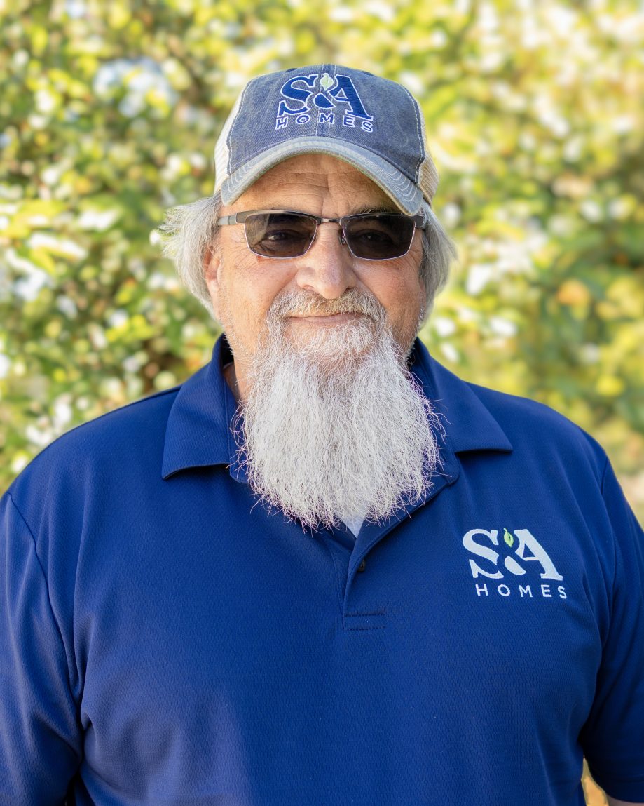 Professional headshot of an S&A Homes team member wearing an S&A Homes polo shirt, cap, and sunglasses, smiling outdoors with trees in the background.