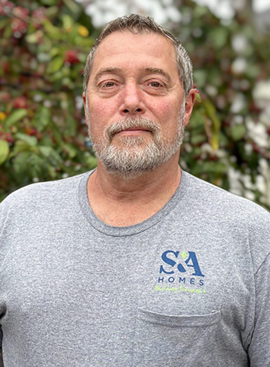 Professional headshot of an S&A Homes team member wearing a gray S&A Homes T-shirt, standing outdoors with green foliage in the background.