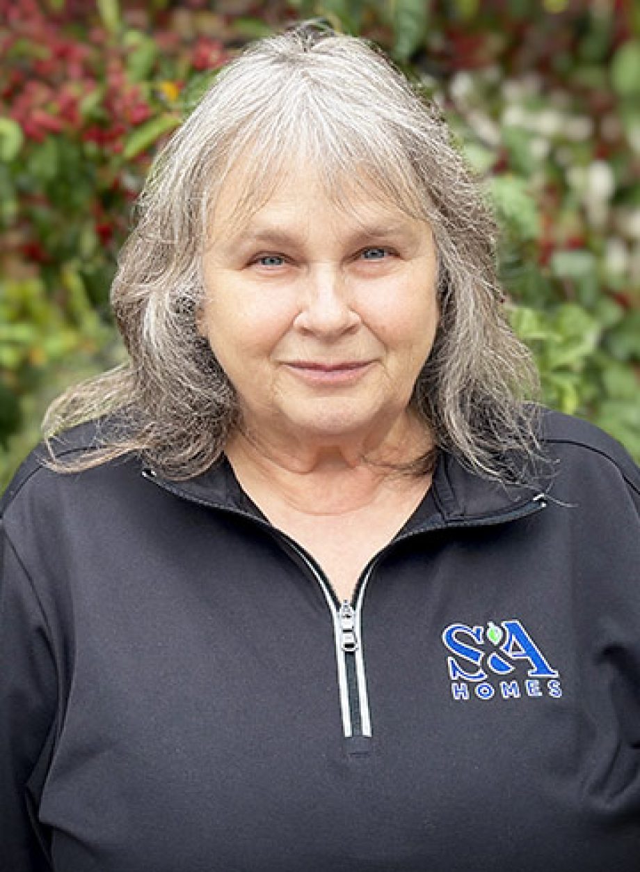 Professional headshot of an S&A Homes team member wearing an S&A Homes quarter-zip pullover, smiling outdoors with green foliage in the background.