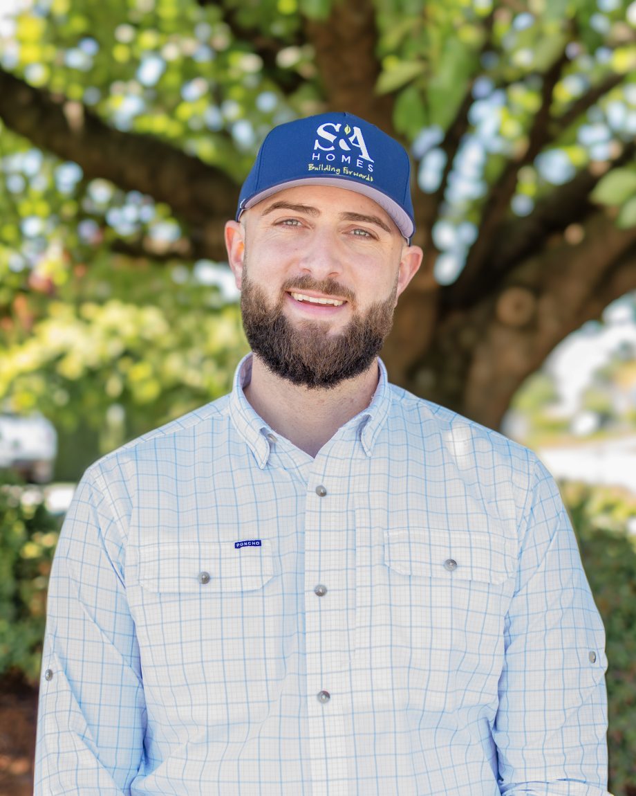 Professional headshot of an S&A Homes team member wearing a polo shirt, cap, and smiling outdoors with trees in the background.