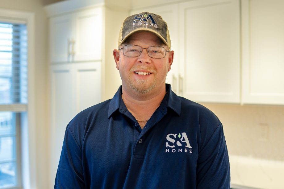 Professional headshot of an S&A Homes team member wearing glasses, an S&A Homes polo shirt, and a baseball cap, smiling in a model home kitchen.